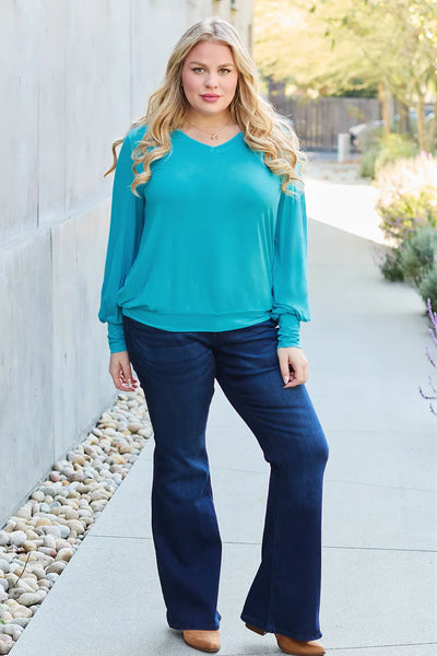 A woman wears a teal v-neck lantern sleeve top and dark wash flare jeans.