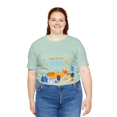 A woman wears a light green t-shirt with a Hanukkah graphic featuring a menorah, dreidels, gelt, and gifts.