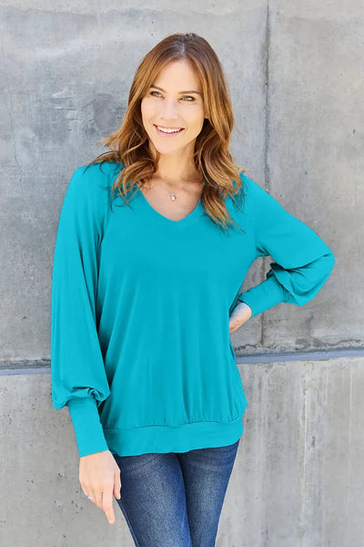 A woman wears a teal v-neck lantern sleeve top and blue jeans.