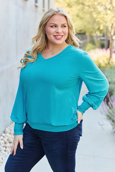 A woman wears a teal v-neck top with lantern sleeves and dark wash jeans.