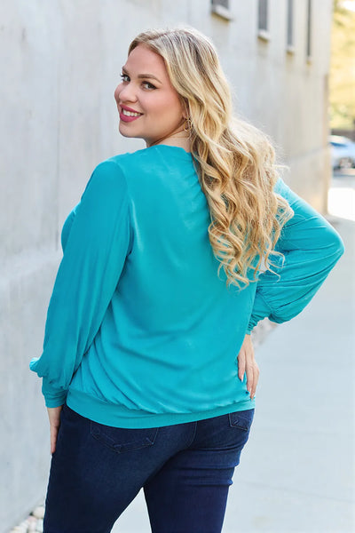 A woman wears a teal V-neck lantern sleeve top and dark wash jeans.